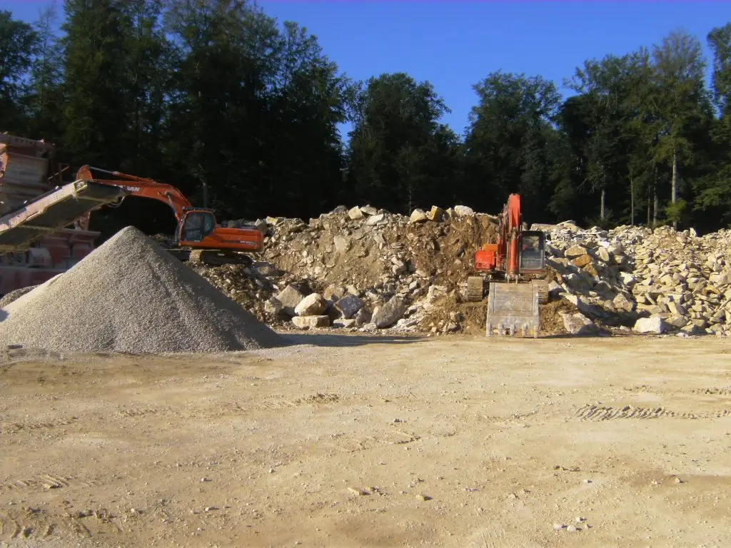 Bschor GmbH: Zwei Bagger arbeiten auf einer Baustelle, der eine neben einem Schotterhaufen, der andere vor einem großen Steinhaufen, mit Bäumen im Hintergrund.
