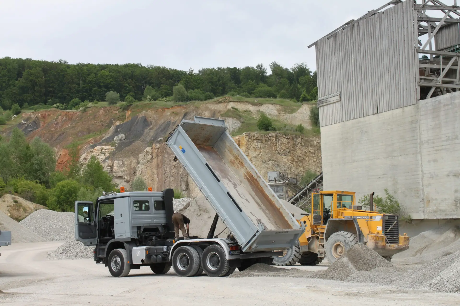 Bschor GmbH: Ein Kipplaster entlädt Kies auf einer Baustelle neben einem Frontlader und einem großen Gebäude, mit einem felsigen Hügel und Bäumen im Hintergrund.
