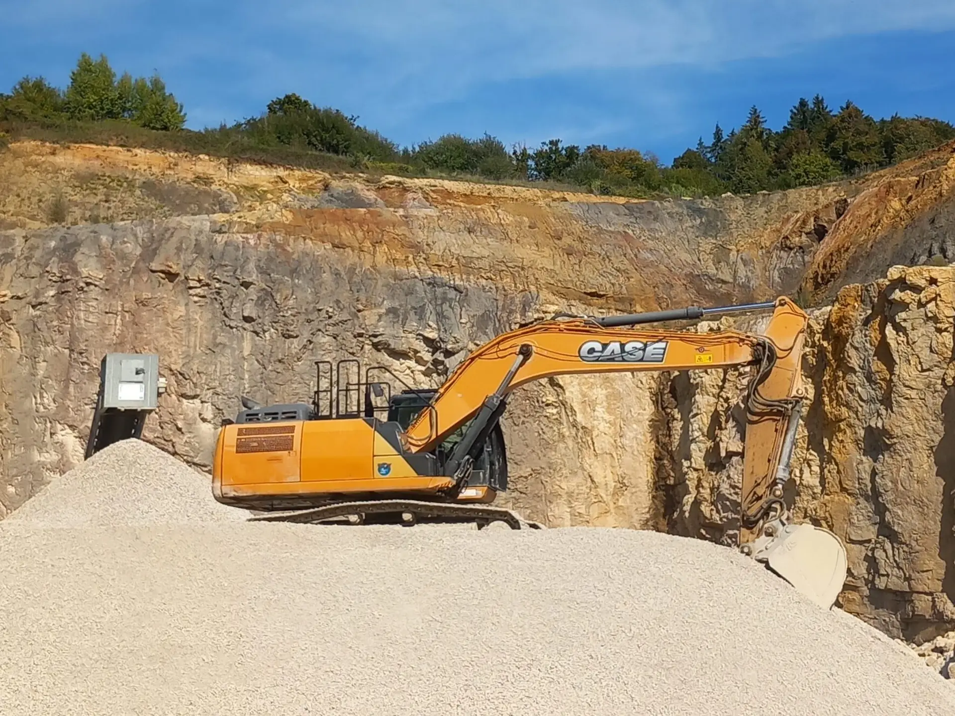 Bschor GmbH: Ein gelber CASE-Bagger ist auf einem Kieshügel in einem Steinbruch geparkt, mit steilen Felswänden und Bäumen im Hintergrund unter einem klaren Himmel.