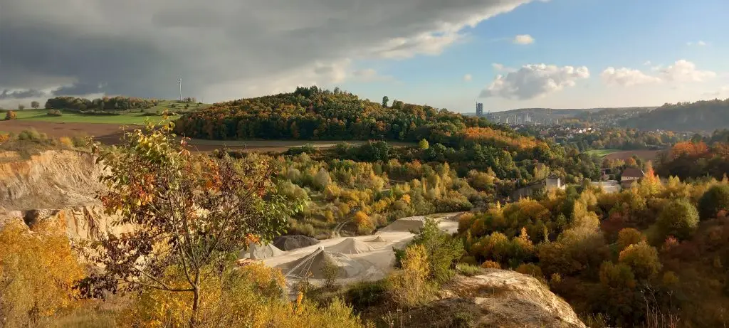 Bschor GmbH: Panoramablick auf eine grüne, hügelige Landschaft mit Herbstlaub, Sand- oder Kiesgruben im Vordergrund, bewölktem Himmel auf der linken Seite und einer entfernten Stadtsilhouette unter blauem Himmel auf der rechten Seite.