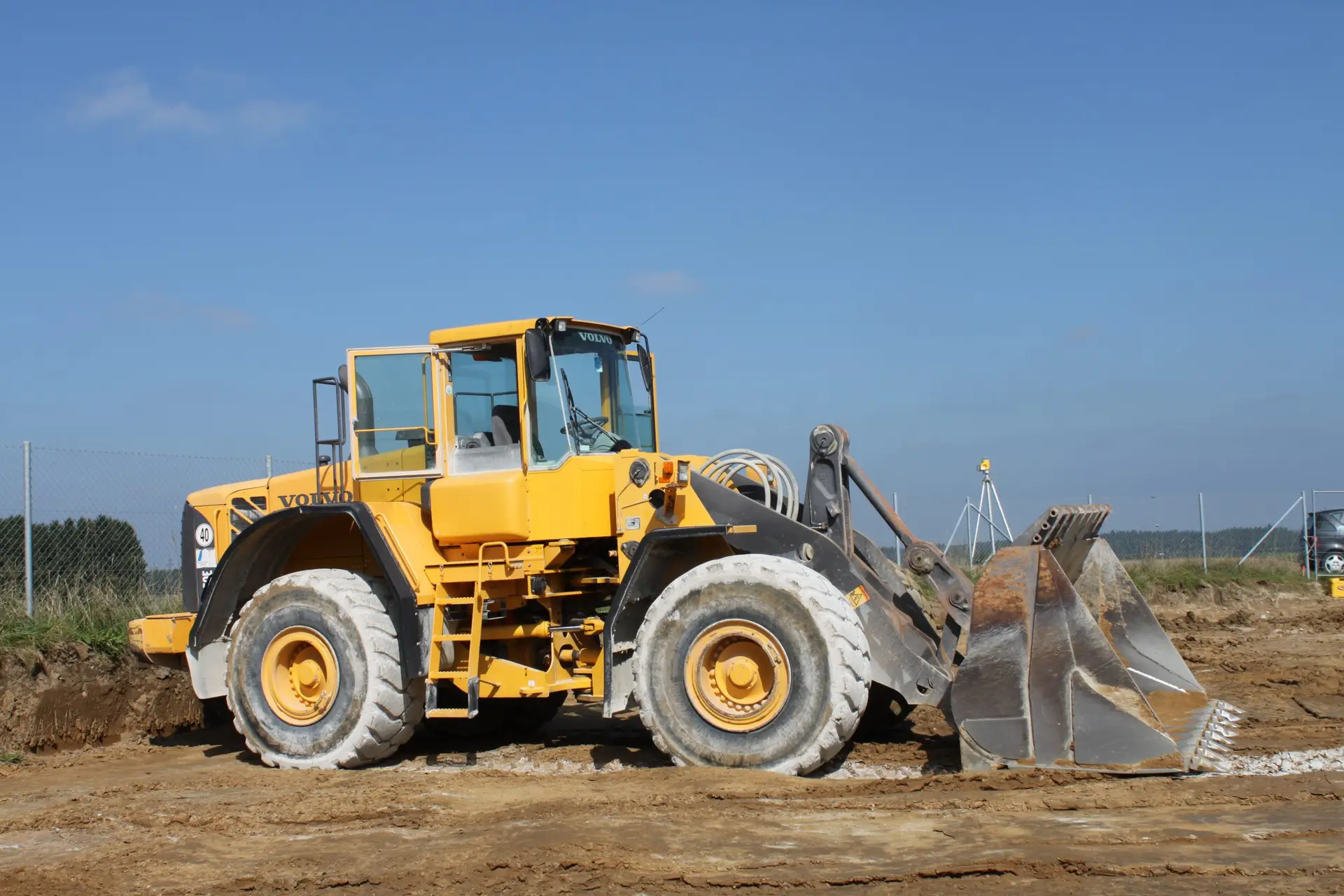 Bschor GmbH: Ein gelber Radlader mit einer großen Frontschaufel ist auf einer Baustelle mit einer unbefestigten Fläche und einem Maschendrahtzaun im Hintergrund abgestellt.