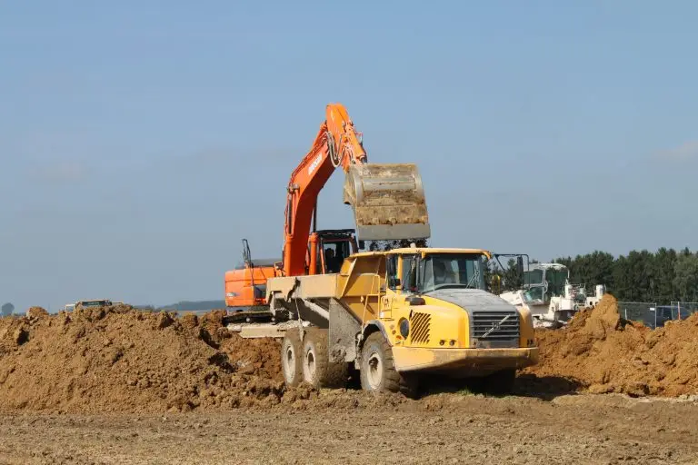 Bschor GmbH: Ein orangefarbener Bagger lädt auf einer Baustelle Erde in einen gelben Muldenkipper, während im Hintergrund bei klarem Himmel Erdhaufen und Baufahrzeuge zu sehen sind.