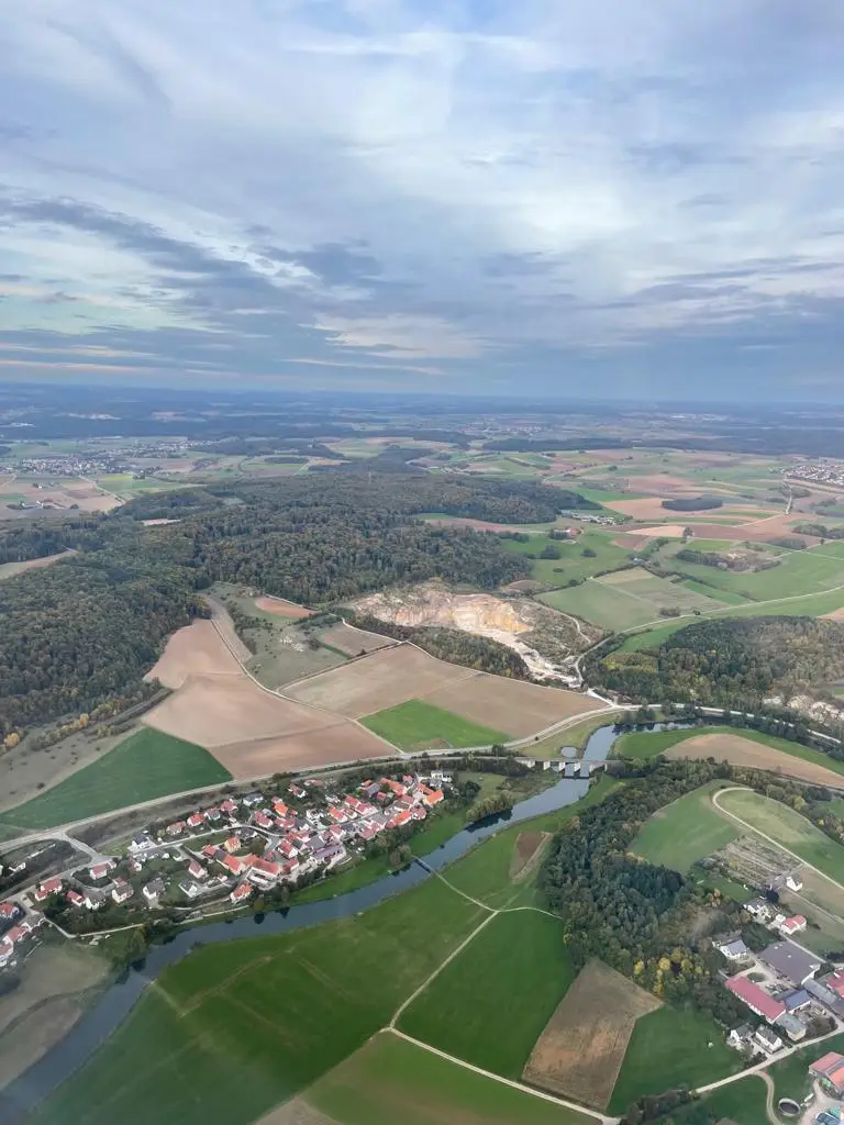 Bschor GmbH: Luftaufnahme einer ländlichen Landschaft mit Feldern, einem kleinen Dorf namens Ronheim, einem gewundenen Fluss und Waldstücken unter einem bewölkten Himmel.