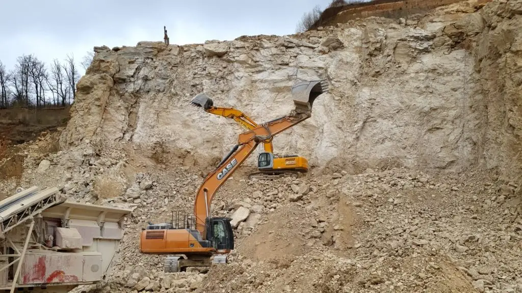 Bschor GmbH: Ein Bagger, der auf felsigem Gelände steht, stützt mit seiner Schaufel einen zweiten Bagger ab, der über dem Boden an einem Steinfelsen steht.