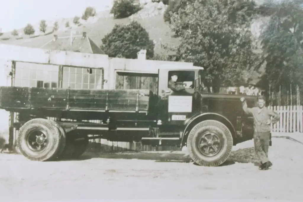 Bschor GmbH: Schwarz-Weiß-Foto eines alten Pritschenwagens, der vor einem Gebäude geparkt ist, wobei ein Mann neben der Fahrertür steht.