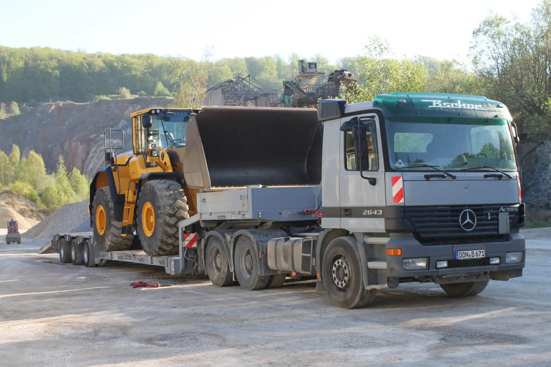 Bschor GmbH: Ein Pritschenwagen transportiert einen großen gelben Radlader auf einem Anhänger auf einer Baustelle im Freien oder in einem Steinbruch.