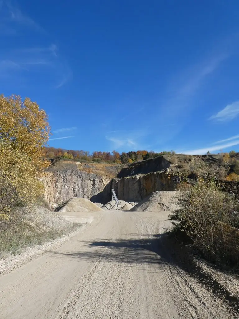 Bschor GmbH: Eine Schotterstraße führt zu einem Steinbruch mit Sand- und Kieshaufen, umgeben von herbstlichen Bäumen und einem strahlend blauen Himmel.