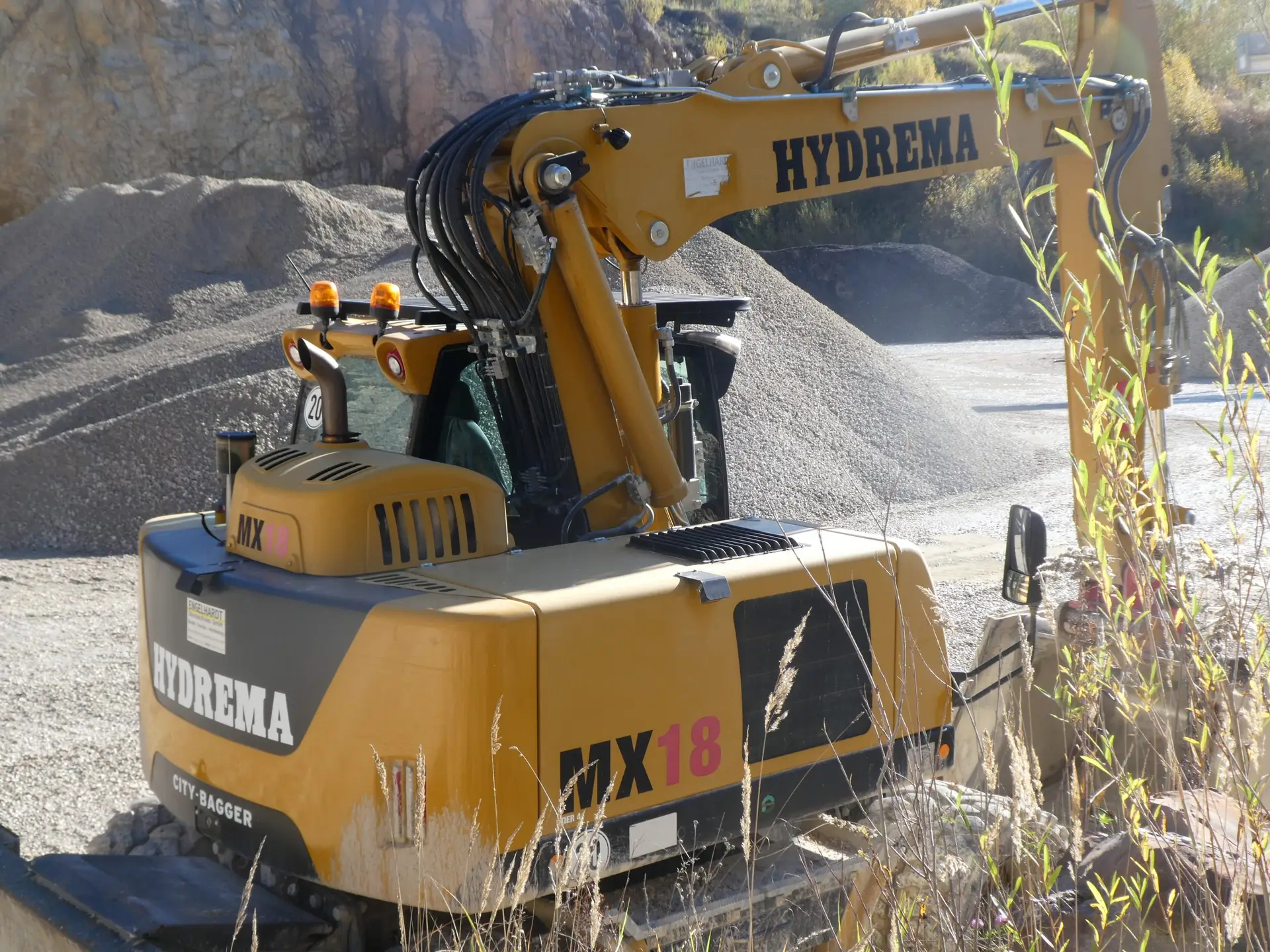 Bschor GmbH: Ein gelber Hydrema MX18-Bagger steht mit teilweise ausgefahrenem Arm auf einer Schotterfläche in der Nähe von Felsenstapeln.