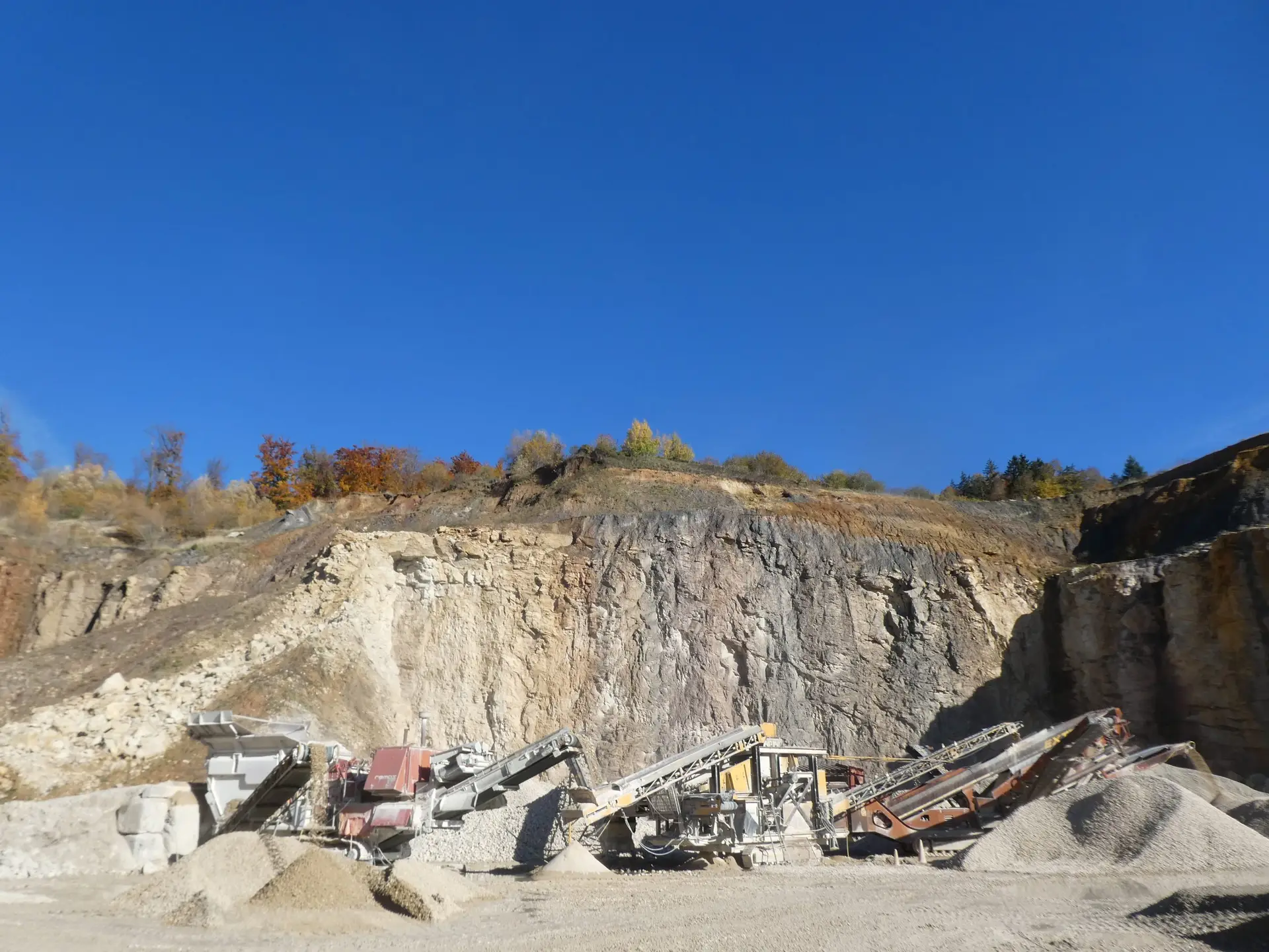 Bschor GmbH: Industrielle Steinbrech- und Sortiermaschinen arbeiten am Fuße einer felsigen Klippe, umgeben von Schotterhaufen unter einem strahlend blauen Himmel.
