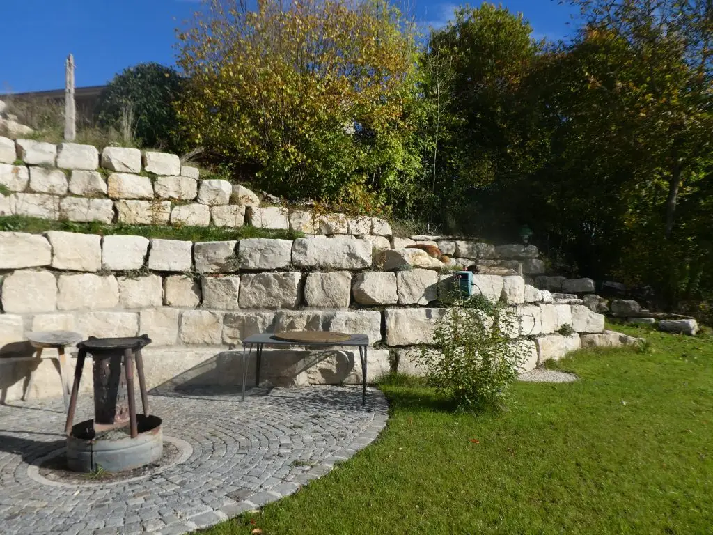 Bschor GmbH: Terrassenförmig angelegte Steinmauern mit grünen Sträuchern und Bäumen, eine Metallbank, eine Feuerstelle und ein runder gepflasterter Bereich auf einer Rasenfläche.