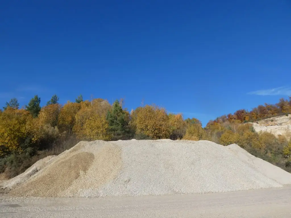 Bschor GmbH: Ein großer Haufen Schottergemisch liegt vor herbstlich gefärbten Bäumen unter einem klaren blauen Himmel.
