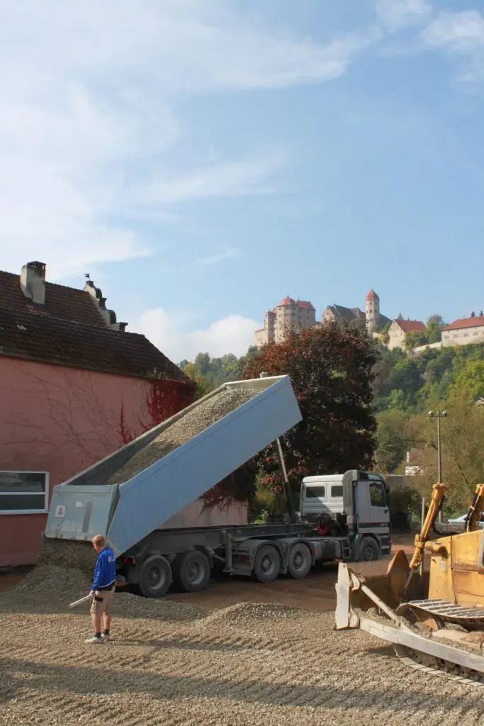 Bschor GmbH: Ein Kipplaster entlädt Kies in der Nähe eines rosafarbenen Gebäudes, während eine Person in blauen Shorts und blauem Hemd in der Nähe auf einer Baustelle steht. Im Hintergrund ist eine Stadt in den Bergen zu sehen.