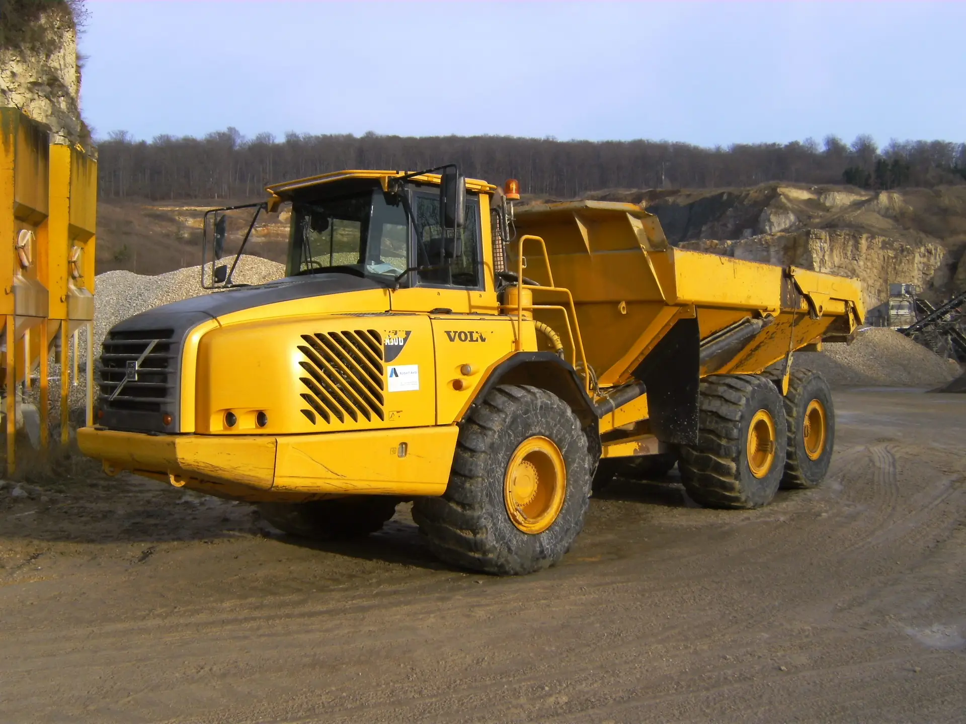 Bschor GmbH: Ein großer gelber knickgelenkter Muldenkipper ist auf einer Kiesfläche in einem Steinbruch geparkt, mit felsigem Gelände im Hintergrund.
