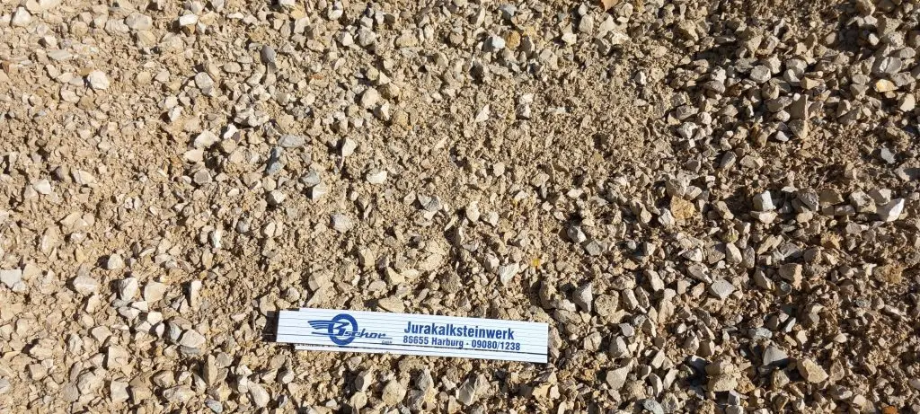 Bschor GmbH: Ein mit Jurakalksteinwerk bezeichnetes Lineal liegt auf einer kiesigen, sandigen Oberfläche, die kleine Steine und eine raue Struktur aufweist.
