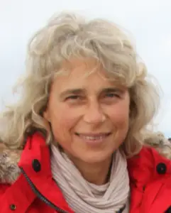 Bschor GmbH: Eine Person mit gewelltem, hellem Haar trägt einen roten Mantel mit pelzbesetzter Kapuze und einen beigen Schal und lächelt in die Kamera vor einem hellen Hintergrund.