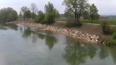Bschor GmbH: Ein ruhiger Fluss fließt entlang einer mit Felsen und Bäumen gesäumten Schotterstraße, mit grünen Feldern und bedecktem Himmel im Hintergrund.
