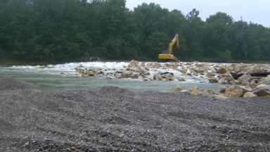 Bschor GmbH: Ein gelber Bagger steht auf Felsen in einem seichten Fluss in der Nähe eines Waldgebiets, mit Kies und Steinen im Vordergrund.