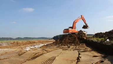Bschor GmbH: Ein orangefarbener Bagger bewegt auf einer Baustelle unter strahlend blauem Himmel einen Erdhaufen, wobei Reifenspuren im sandigen Boden sichtbar sind.
