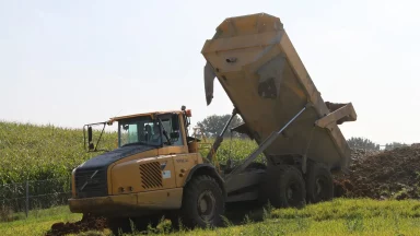 Bschor GmbH: Ein gelber Muldenkipper entlädt an einem sonnigen Tag Erde auf einer Wiese in der Nähe eines Maisfeldes.