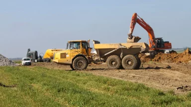 Bschor GmbH: Ein gelber Kipper und ein orangefarbener Bagger arbeiten auf einer Baustelle und bewegen unter freiem Himmel Erde in der Nähe einer Grasfläche.