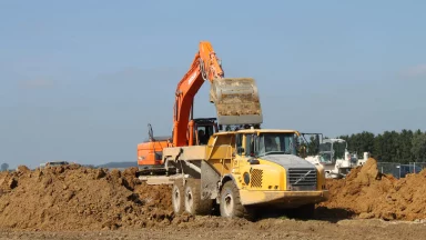 Bschor GmbH: Ein orangefarbener Bagger lädt auf einer Baustelle Erde in einen gelben Muldenkipper, während im Hintergrund bei klarem Himmel Erdhaufen und Baufahrzeuge zu sehen sind.