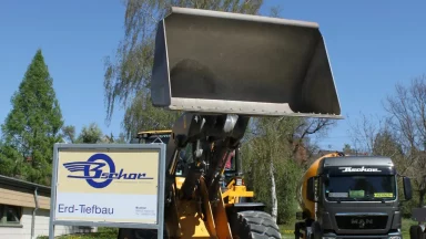 Bschor GmbH: Ein großer gelber Frontlader mit angehobener Schaufel ist neben einem Firmenschild und einem Zementmischer in einem sonnigen Außenbereich abgestellt.