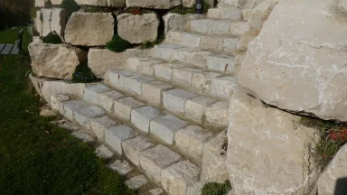 Bschor GmbH: Eine Steintreppe aus großen, grob behauenen Kalksteinblöcken führt einen kleinen Hang hinauf, der von Grün und Gras gesäumt wird.