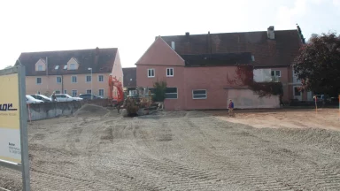 Bschor GmbH: Eine Baustelle mit Schotterboden vor Wohngebäuden, mit einem Bulldozer, einem Bagger und einer Person, die in der Nähe geht.