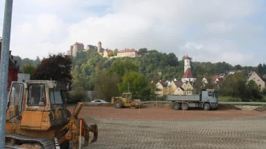 Bschor GmbH: Baufahrzeuge auf einer geräumten Baustelle im Vordergrund; im Hintergrund sind bei teilweise bewölktem Himmel eine Burg auf einem Hügel und städtische Gebäude zu sehen.
